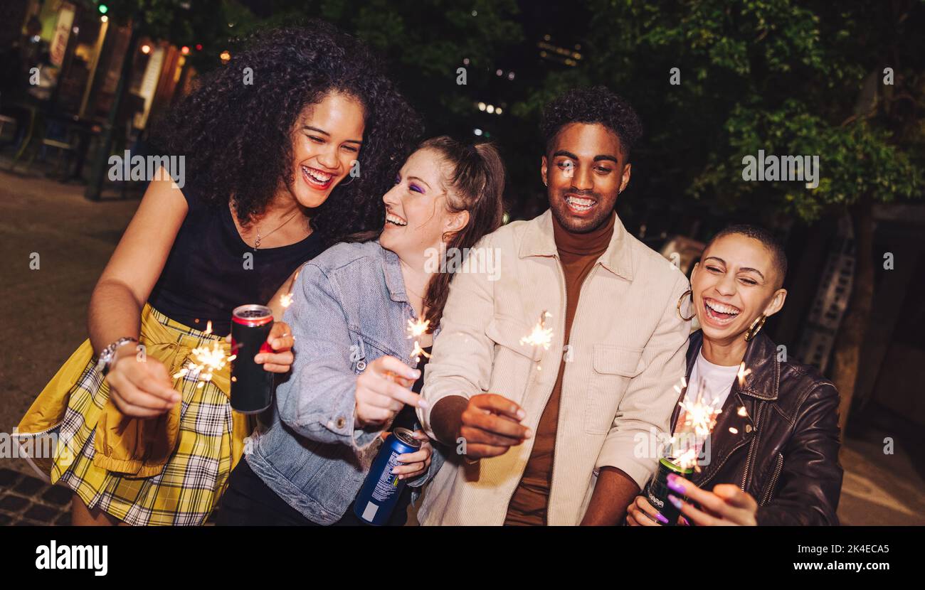 Amici della città che festeggiano la stagione delle feste con gli scintillanti. Gruppo di giovani felici che tengono le luci del bengala e lattine di birra di notte. Amici allegri Foto Stock