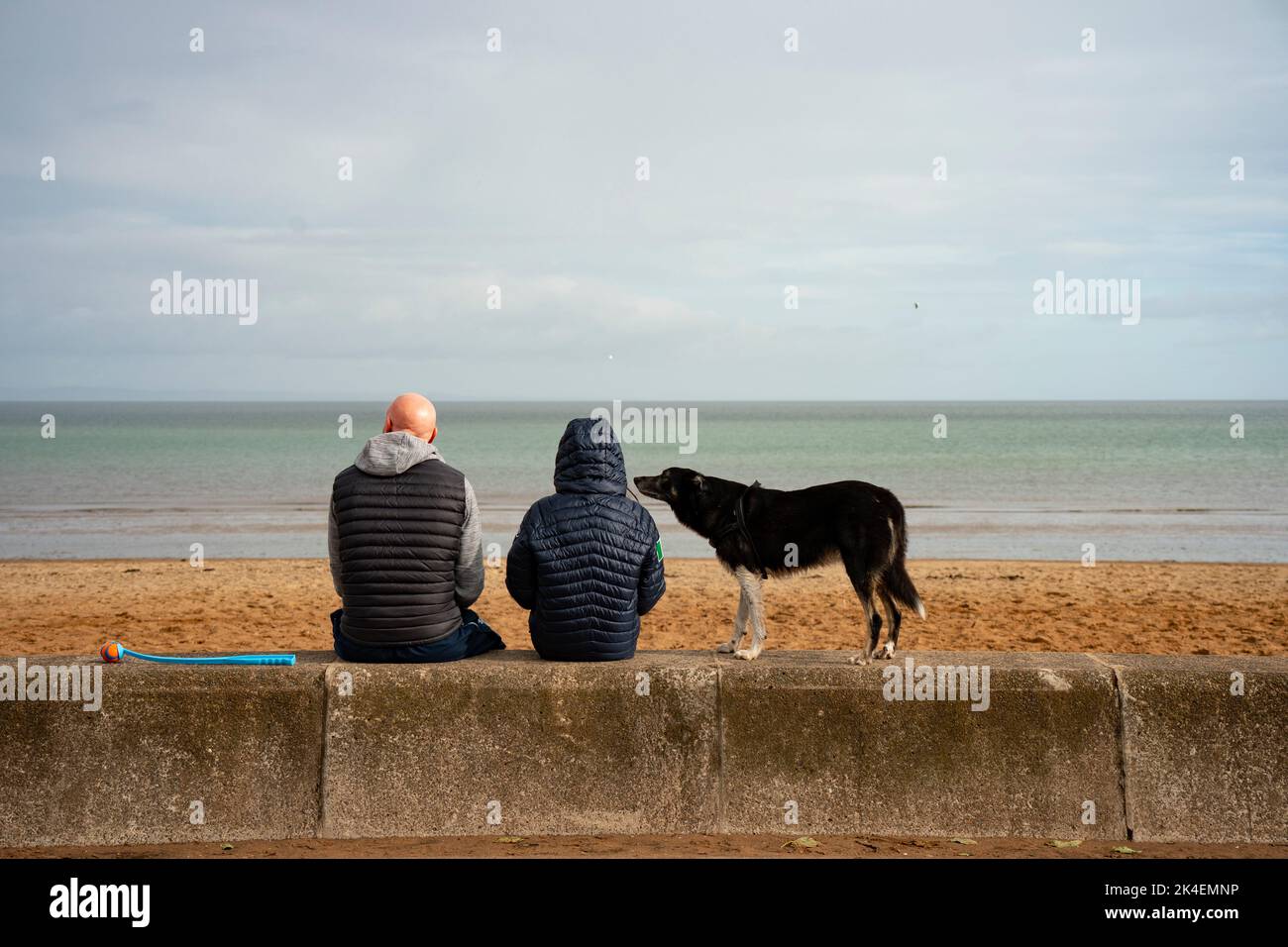 Coppia seduta sul muro di mare accanto alla spiaggia con cane da compagnia a Portobello, Edimburgo, Scozia, Regno Unito Foto Stock