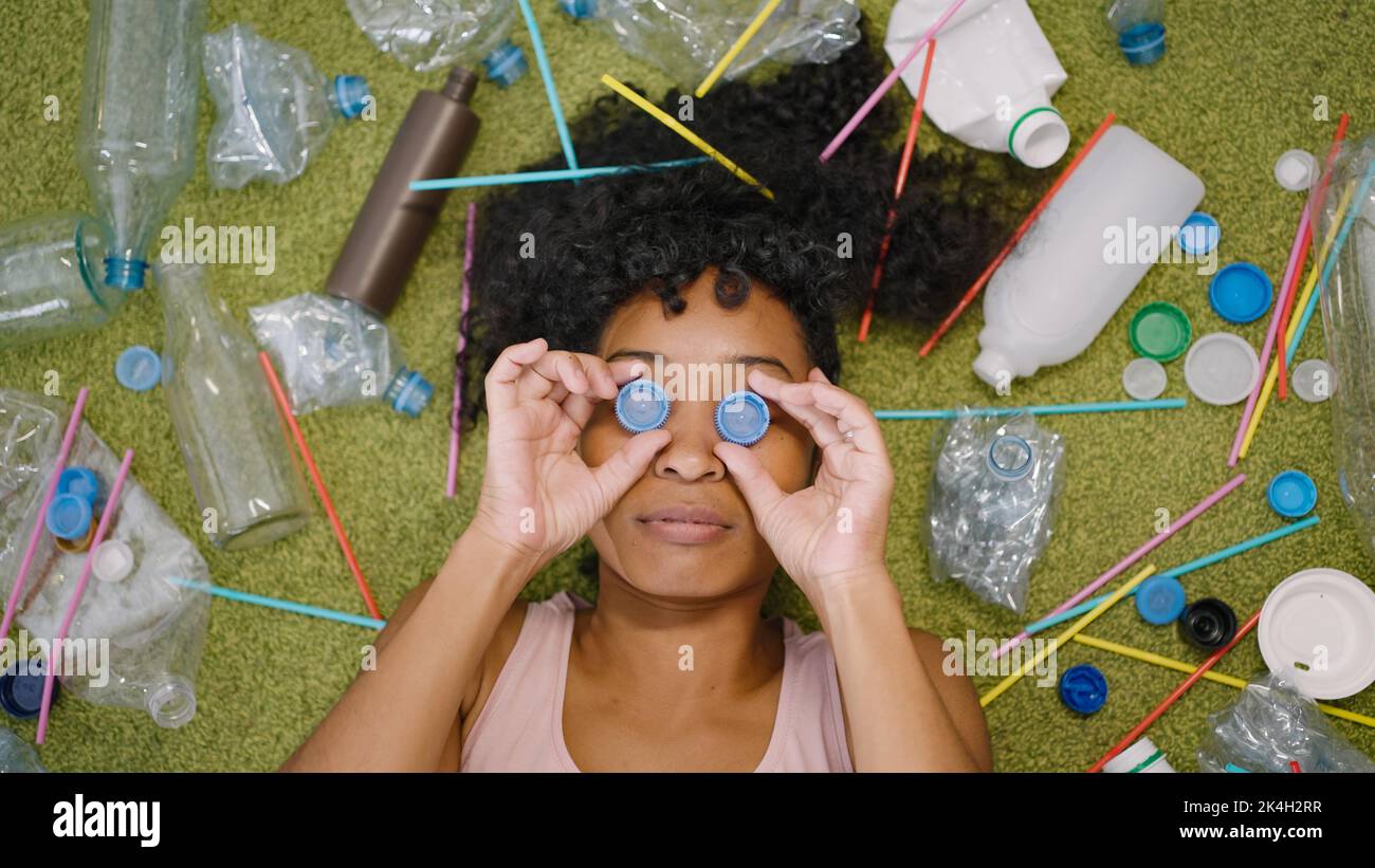 La donna afroamericana mette i tappi di plastica dalle bottiglie sugli occhi. Casalinga si trova su coperta vicino spazzatura sparsa che enfatizza il problema di primo piano superiore Foto Stock
