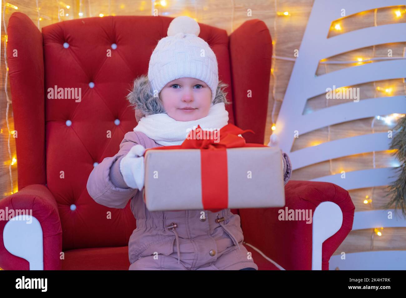 Ritratto bambina che si siede in una sedia rossa di natale e dà un regalo di Natale fuori. Natale presenta il concetto di infanzia di Vacanze Foto Stock
