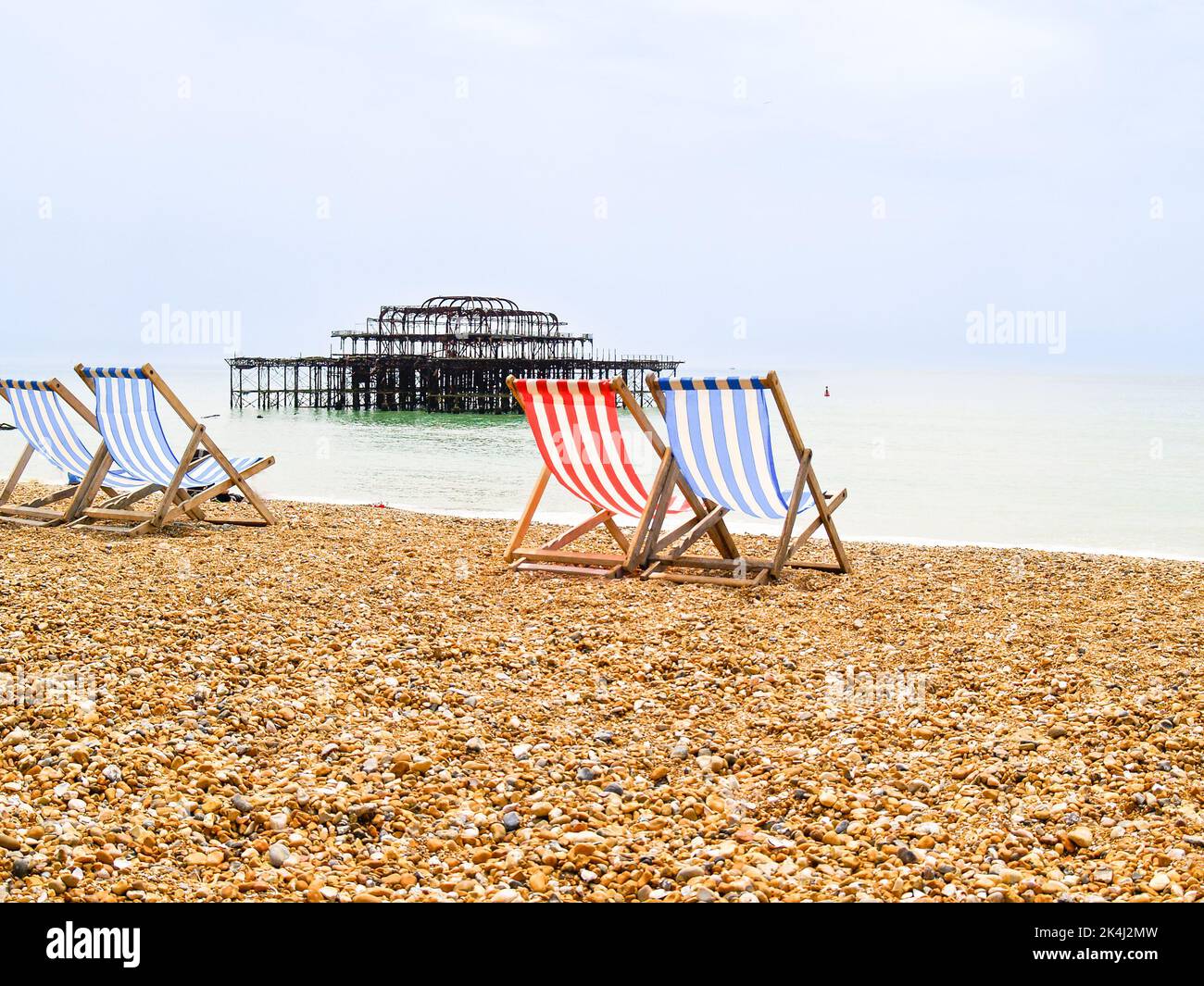 Brighton Beach e le caratteristiche sedie a sdraio a strisce in blu o rosso e bianco. E resti del vecchio Brighton Pier appena fuori dalla riva. Foto Stock