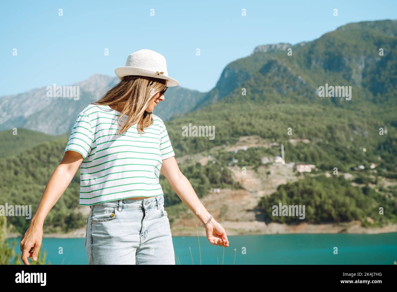 Donna sorridente in cappello e occhiali da sole con capelli selvatici in piedi vicino lago montagne sullo sfondo. Positiva giovane donna che viaggia sul lago blu all'aperto Foto Stock