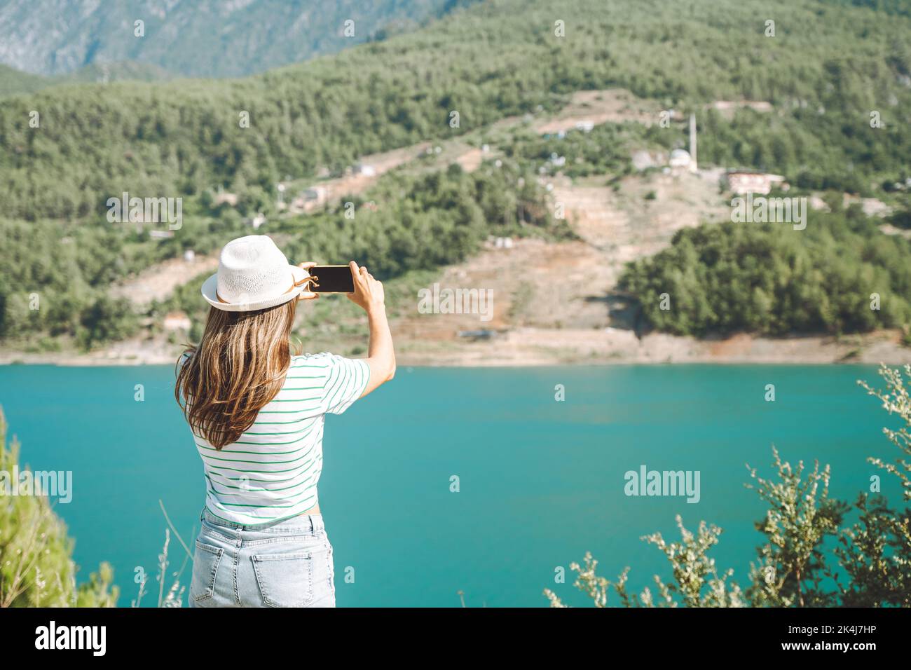 Donna in cappello con capelli selvatici che scatta un video sul telefono cellulare di montagne lago sfondo. Viaggiatore che scatta foto sul cellulare sul lago blu Foto Stock