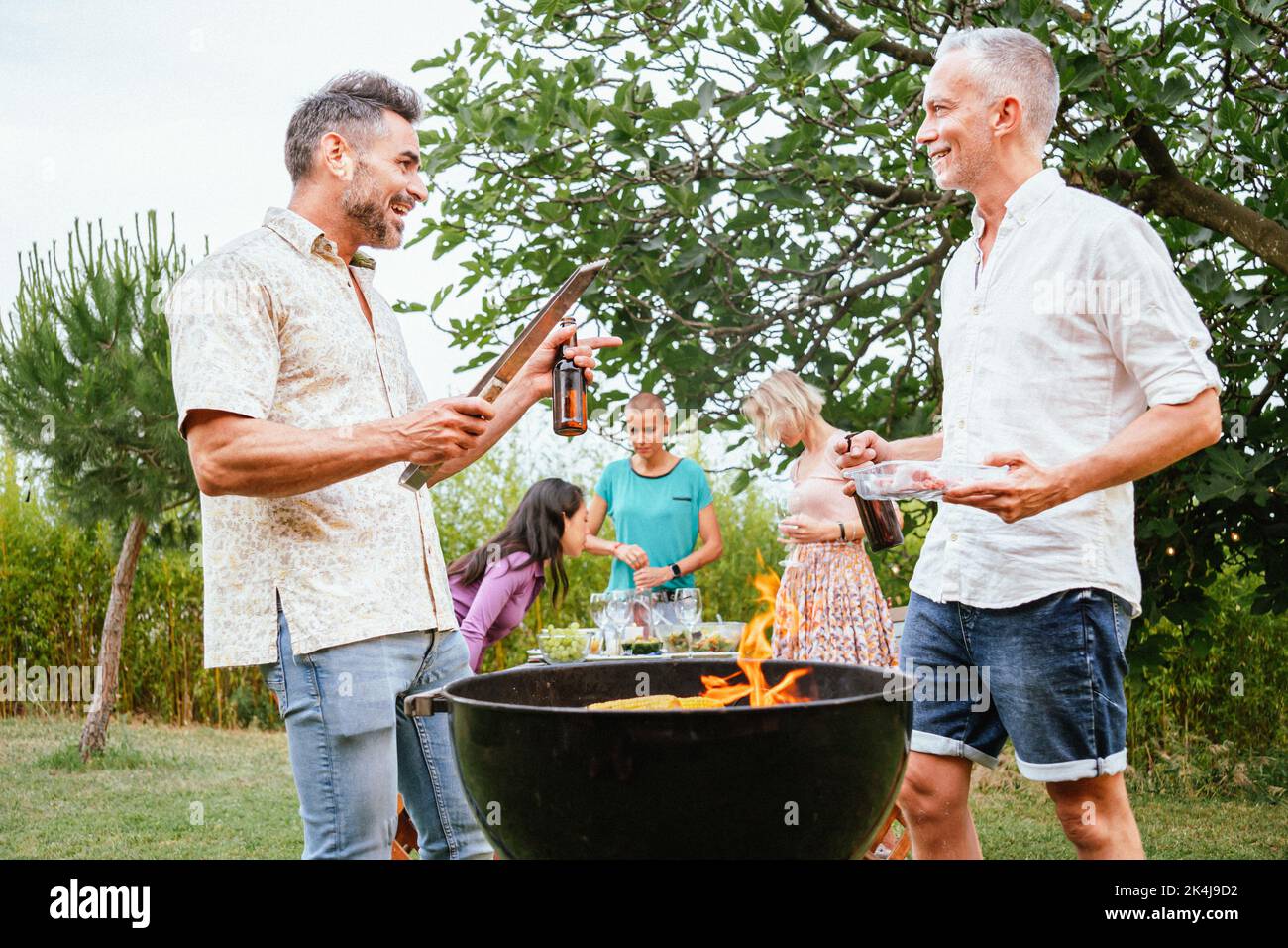 Due amici adulti o uomini di famiglia che parlano e sorridono, cucinano e grigliano in una festa barbecue con i suoi amici multietnici felici. Concetto di stile di vita. Foto Stock