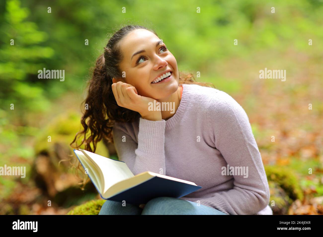 Donna felice sognando mentre legga un libro in una foresta che guarda sopra Foto Stock