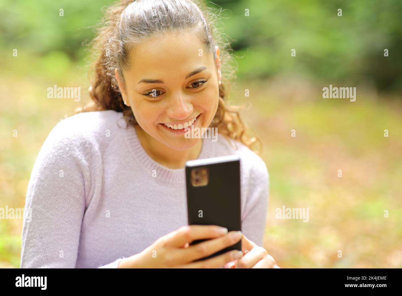 Donna felice seduta in una foresta o in un parco con smartphone Foto Stock