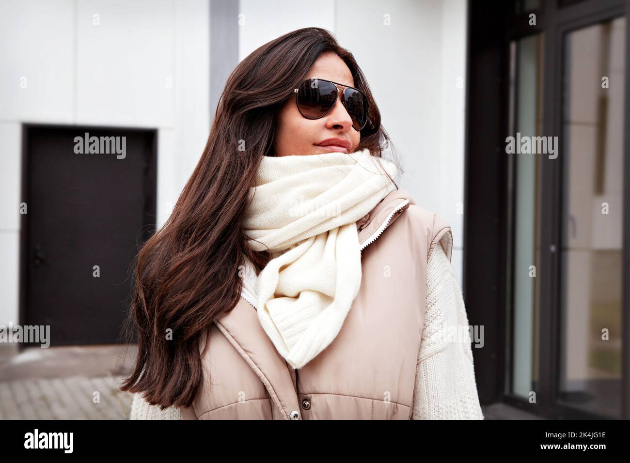 Primo piano Ritratto di bella donna che guarda via sulla strada. Modello elegante con lunghi capelli brunette che indossano occhiali da sole, sciarpa bianca lavorata a maglia e maglione Foto Stock