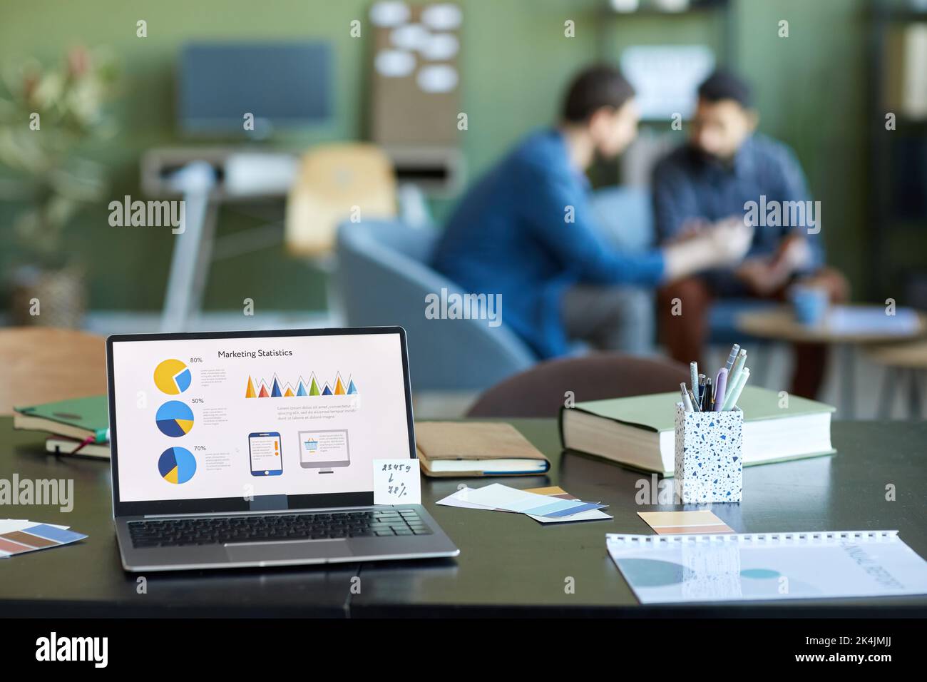 Schermo di un computer portatile con dati finanziari e una varietà di forniture di lavoratori in ufficio sulla scrivania con due uomini d'affari che discutano in background Foto Stock