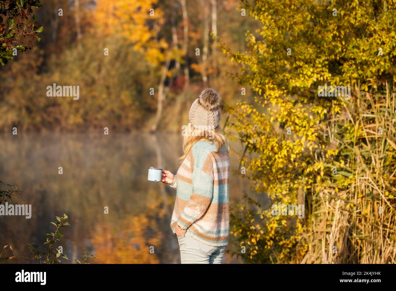 Donna con maglione e cappello in maglia che si rilassa con una bevanda calda accanto al lago durante la fredda mattinata autunnale Foto Stock