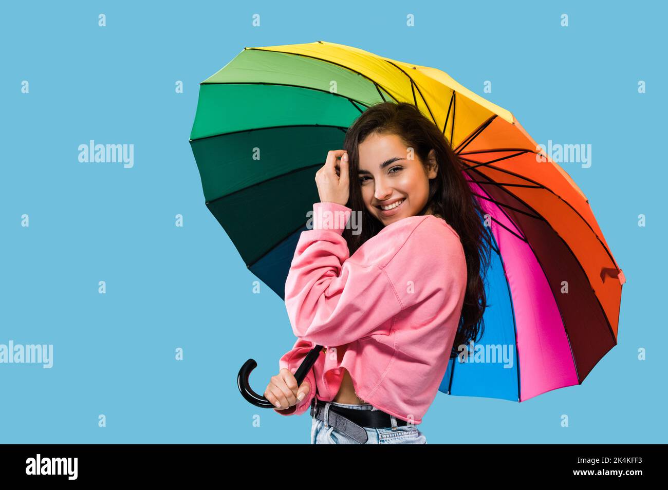 Bella positiva giovane donna caucasica elegante con capelli lunghi, in abbigliamento casual, in piedi su sfondo isolato blu da ombrello arcobaleno colorato, guardando la macchina fotografica, sorridendo felicemente Foto Stock