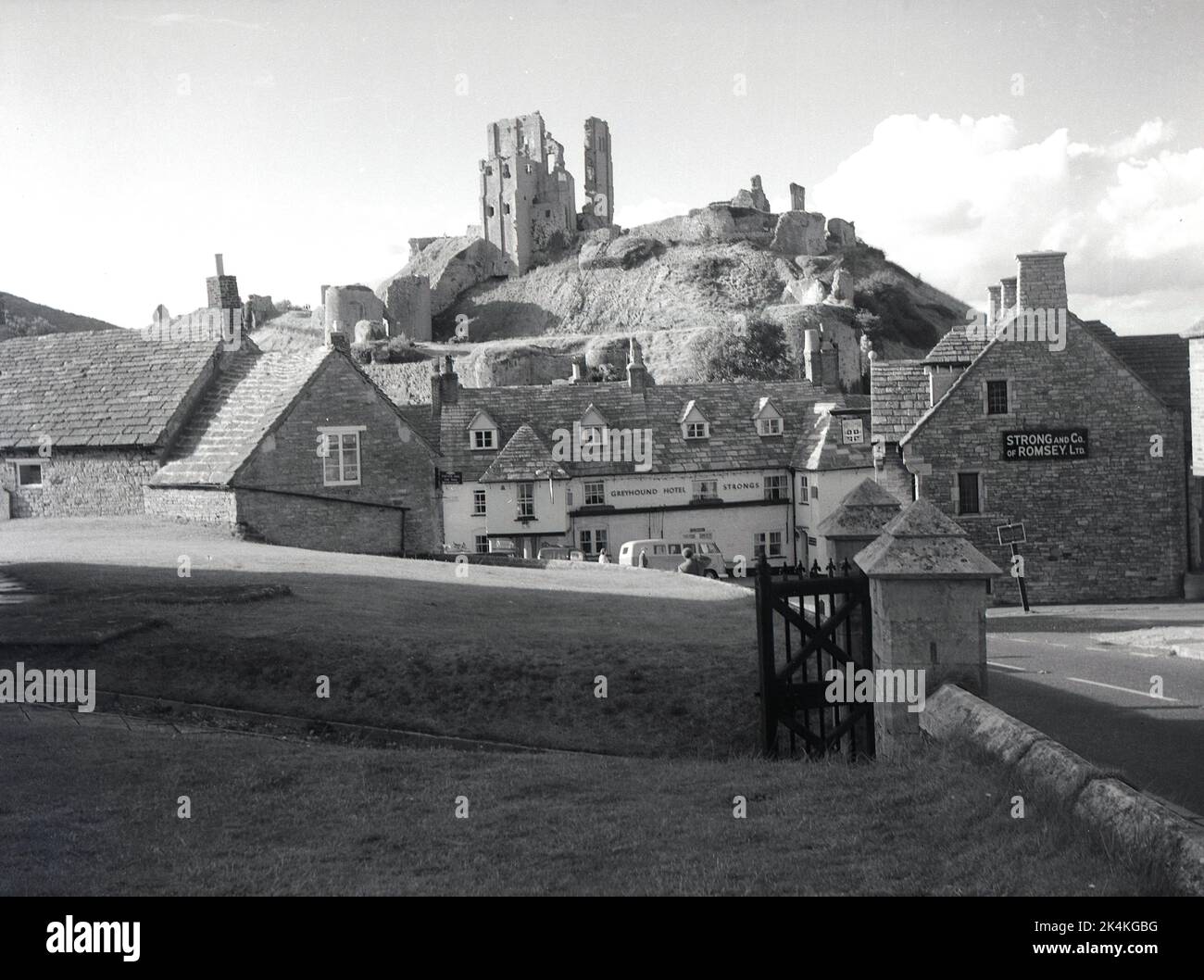 1964, storico, il villaggio di Corfe Caste e una vista del Greyhound Hotel, con le rovine del castello di Corfe in cima alla collina, Dorset, Inghilterra, Regno Unito. Firma per strong and Co of Romsey Ltd sulla parete dell'edificio. In seguito divenne un pub Whitebread. Risalente al 1580, il pub è una delle più antiche pensioni per carrozze d'Inghilterra. Originariamente costruito da Guglielmo il Conquistatore, il castello in cima alla collina era una delle prime fortificazioni costruite in parte in pietra, ma nel 1645 fu leggermente distrutto, cioè parzialmente distrutto, dopo la guerra civile inglese, per evitare che fosse utilizzato come una potenziale fortezza e divenne una rovina. Foto Stock