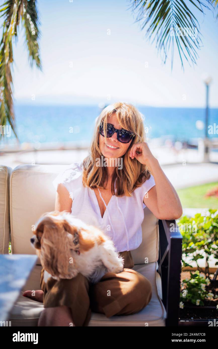 Foto di donna di mezza età godendo di una giornata di sole mentre si rilassa sul mare con il suo cucciolo di cavalier re charles. Foto Stock