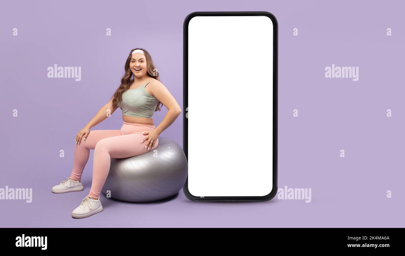 Felice europeo giovane donna più dimensione fare esercizi sulla palla fitness vicino grande smartphone con schermo vuoto Foto Stock