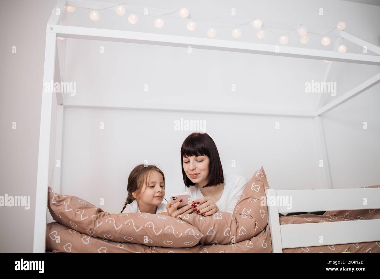 Bella giovane donna e la sua affascinante bambina in auricolari stanno ascoltando la musica utilizzando uno smartphone e sorridendo mentre sdraiato a letto a casa Foto Stock