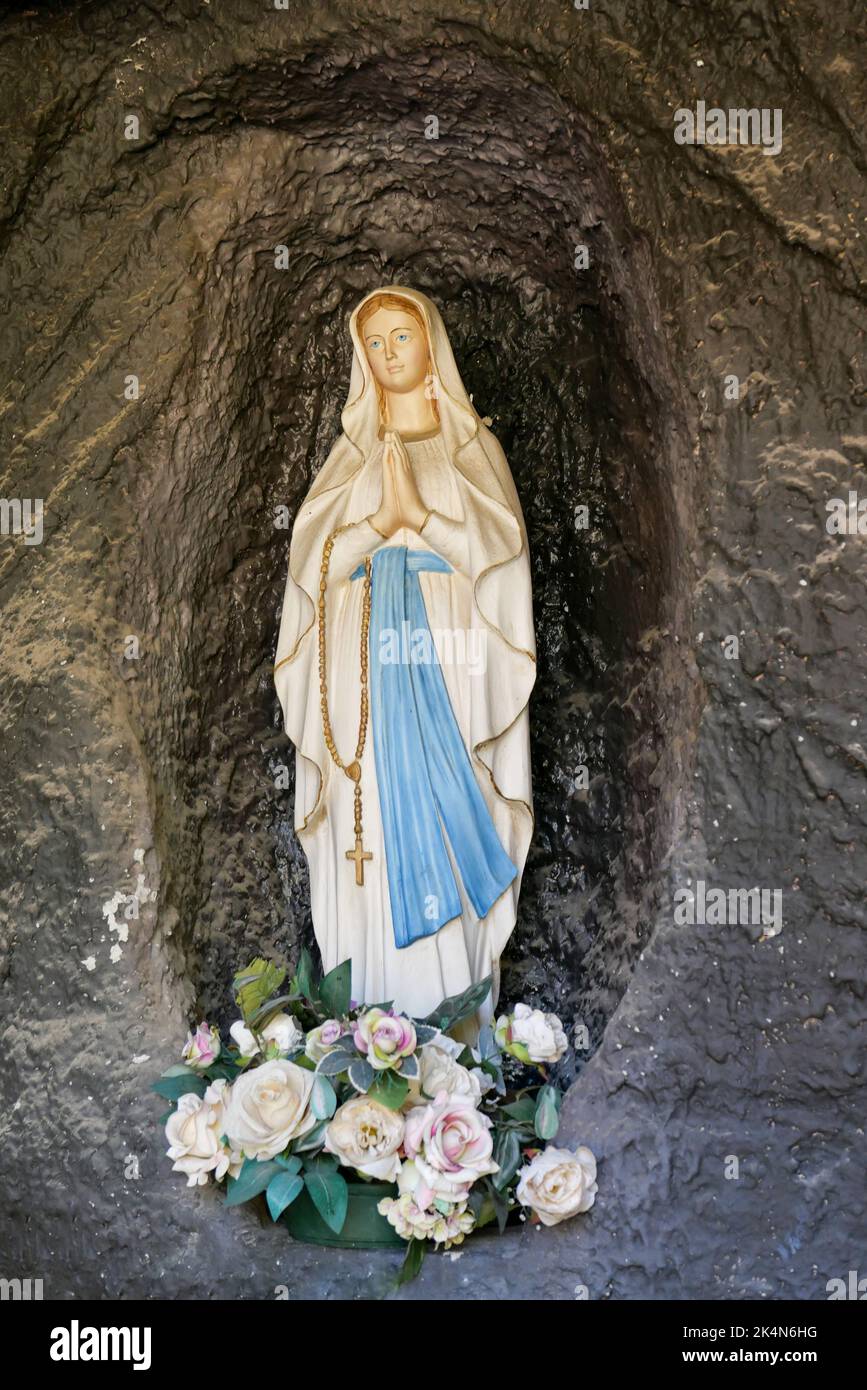 Una vista della statua di Santa Maria che prega in una grotta di pietra con fiori ai suoi piedi Foto Stock