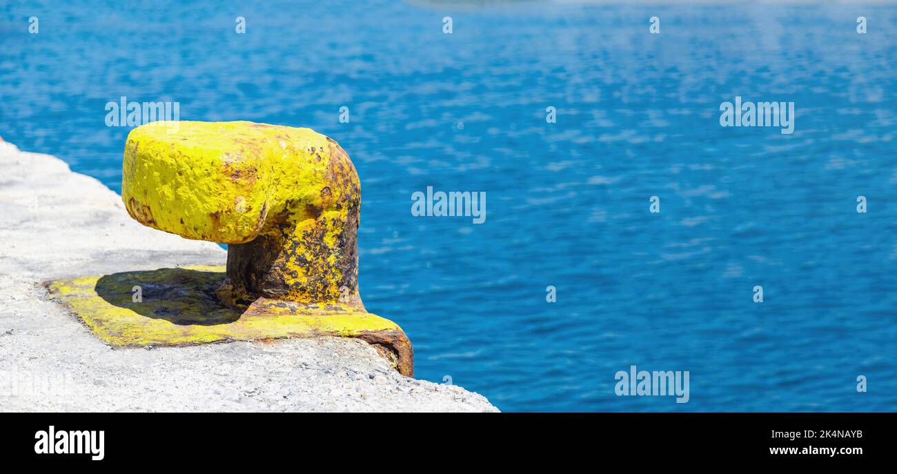 Giallo arrugginito porto bollard sul molo del porto, sfocato acqua di mare sfondo, copia spazio. Vecchio palo di metallo ancorato sul pavimento in cemento della banchina dell'isola greca. Foto Stock