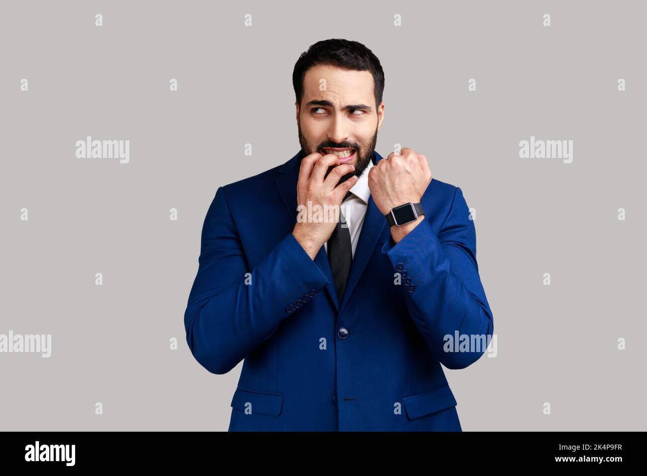 Nervoso uomo bearded mordere unghie e guardare la macchina fotografica, mostrando il suo orologio da polso, preoccupato per la scadenza, indossando abiti in stile ufficiale. Studio in interni isolato su sfondo grigio. Foto Stock
