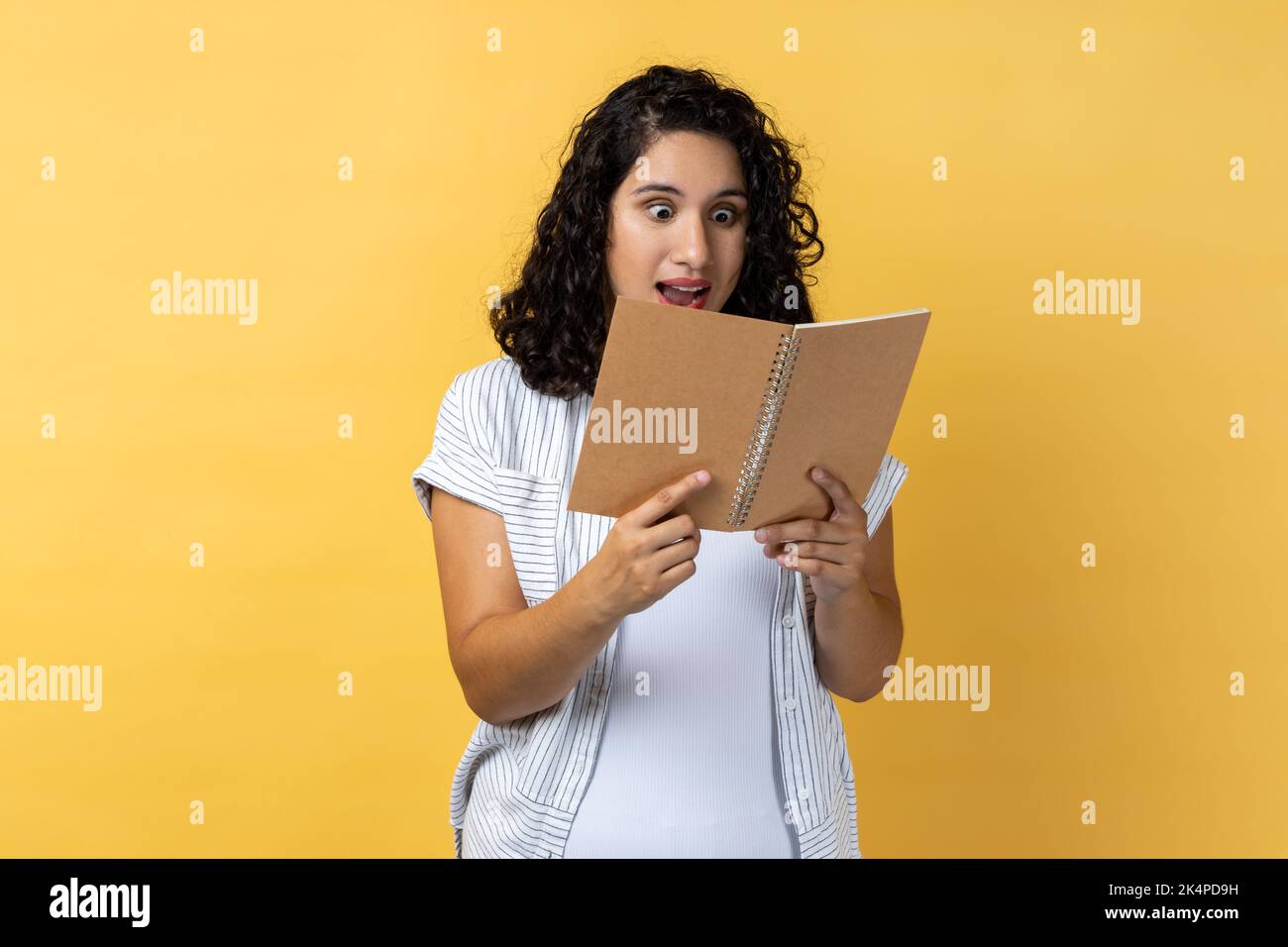 Ritratto di stupito impressionato donna sorpresa con i capelli ondulati scuri in piedi con il libro nelle mani, leggendo romanzo stupefacente, guardando le pagine con gli occhi grandi. Studio al coperto isolato su sfondo giallo. Foto Stock