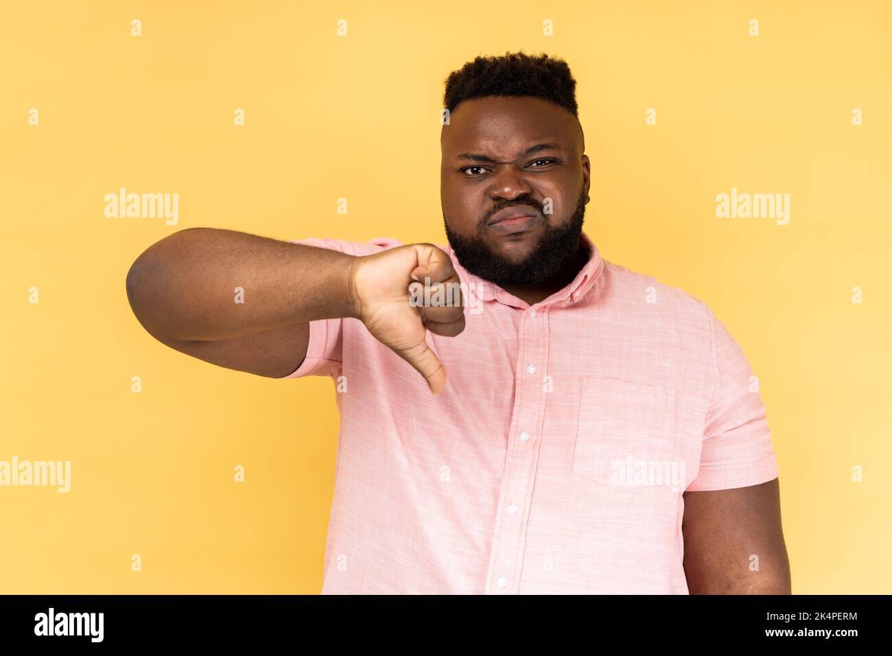 Ritratto di triste sconvolto uomo bearded indossare camicia rosa mostrando il pollice giù, esprimendo le emozioni negative difettose, dislike, faccia accigliante. Studio al coperto isolato su sfondo giallo. Foto Stock