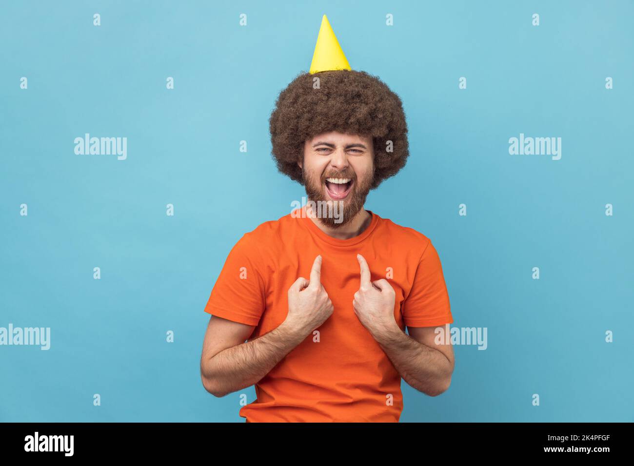 Ritratto di pazzo uomo felice con acconciatura afro indossare T-shirt arancione e cono festa, guardando la macchina fotografica, indicando se stesso, celebrando il suo compleanno. Studio in interni isolato su sfondo blu. Foto Stock