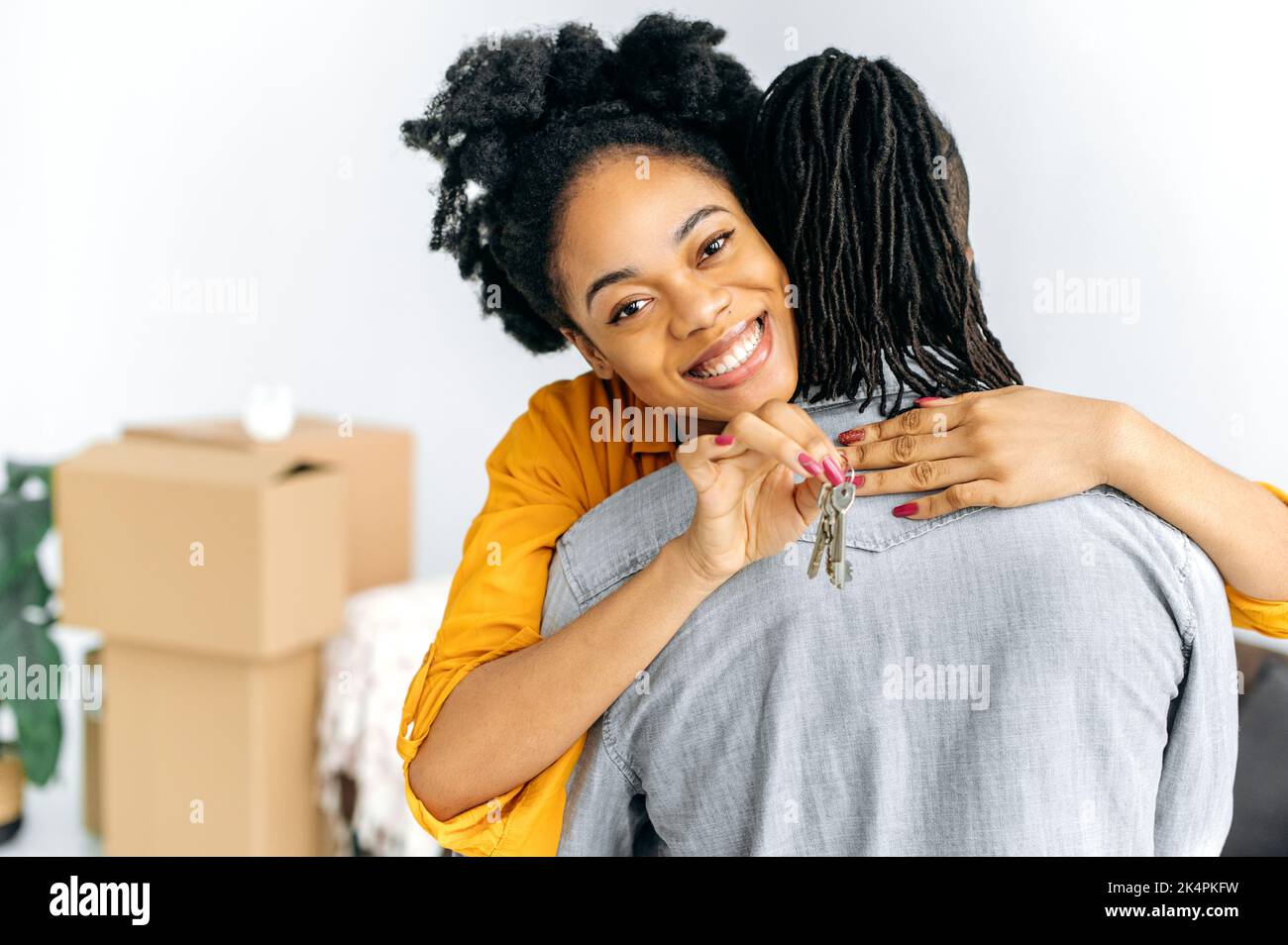 Investimento, concetto di proprietà. Una donna afro-americana eccitata positiva che abbraccia suo marito mentre si trova in piedi in salotto, mostrando le chiavi della loro nuova proprietà, guarda la macchina fotografica, sorridendo felicemente Foto Stock