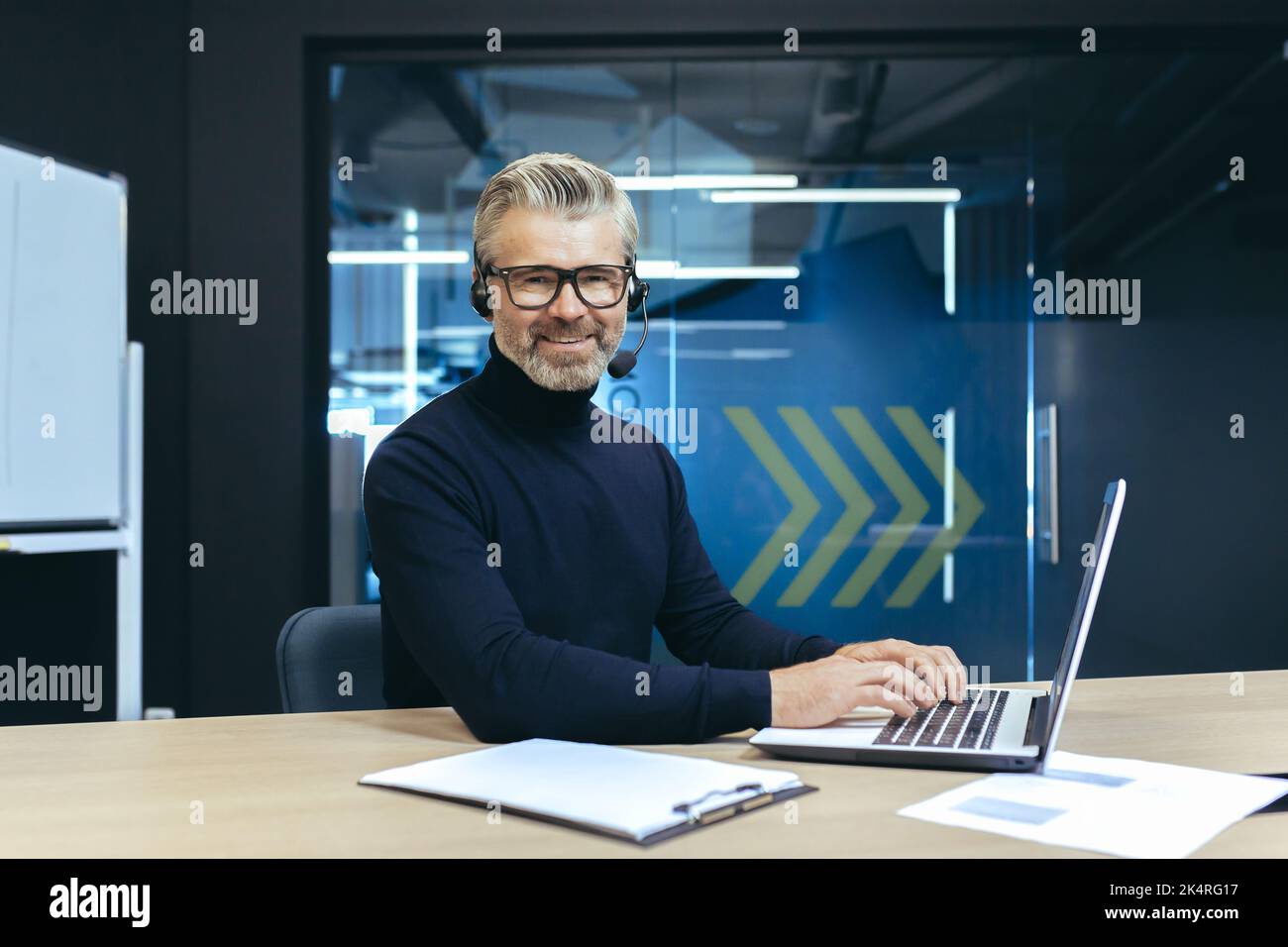 Un bell'uomo d'affari dai capelli grigi con una cuffia e un microfono sta lavorando su un computer portatile. Si siede in ufficio al tavolo, conduce videoconferenze, consulta. Guarda la telecamera, sorride. Foto Stock