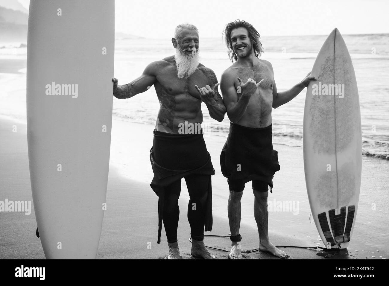 Amici di surfista multigenerazionale che si divertono in spiaggia dopo una sessione di surf - Focus on Faces - editing in bianco e nero. Foto Stock