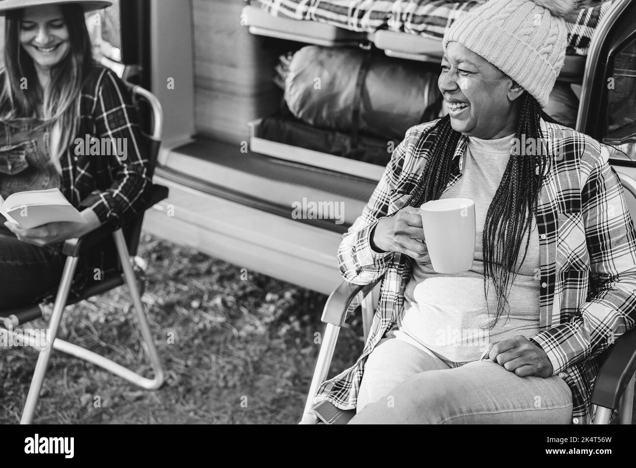 Amici multirazziali donne divertirsi campeggio con camper mentre leggere e bere caffè all'aperto - Focus on african female face - e bianco e nero Foto Stock