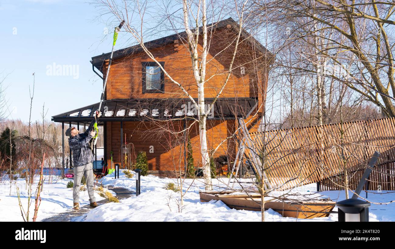Manico telescopico di tagliasiepi. Architettura paesaggistica nella stagione invernale. Un giardiniere pota un albero con una potatrice a catena elettrica nella stagione invernale. Inverno Foto Stock