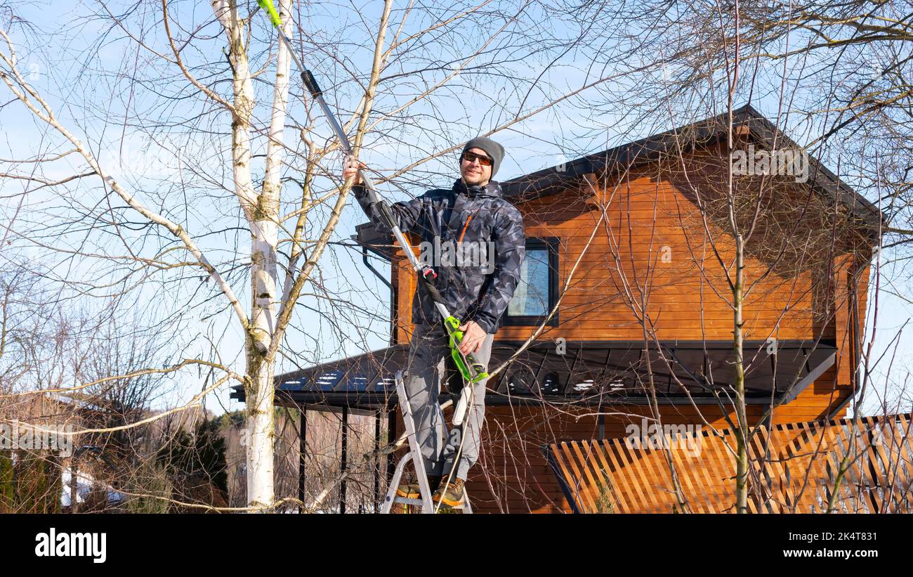 Un giardiniere è in piedi su una scala con una catena elettrica potatrice da giardino con un palo telescopico e il taglio di rami di albero in attrezzatura invernale. Wint Foto Stock