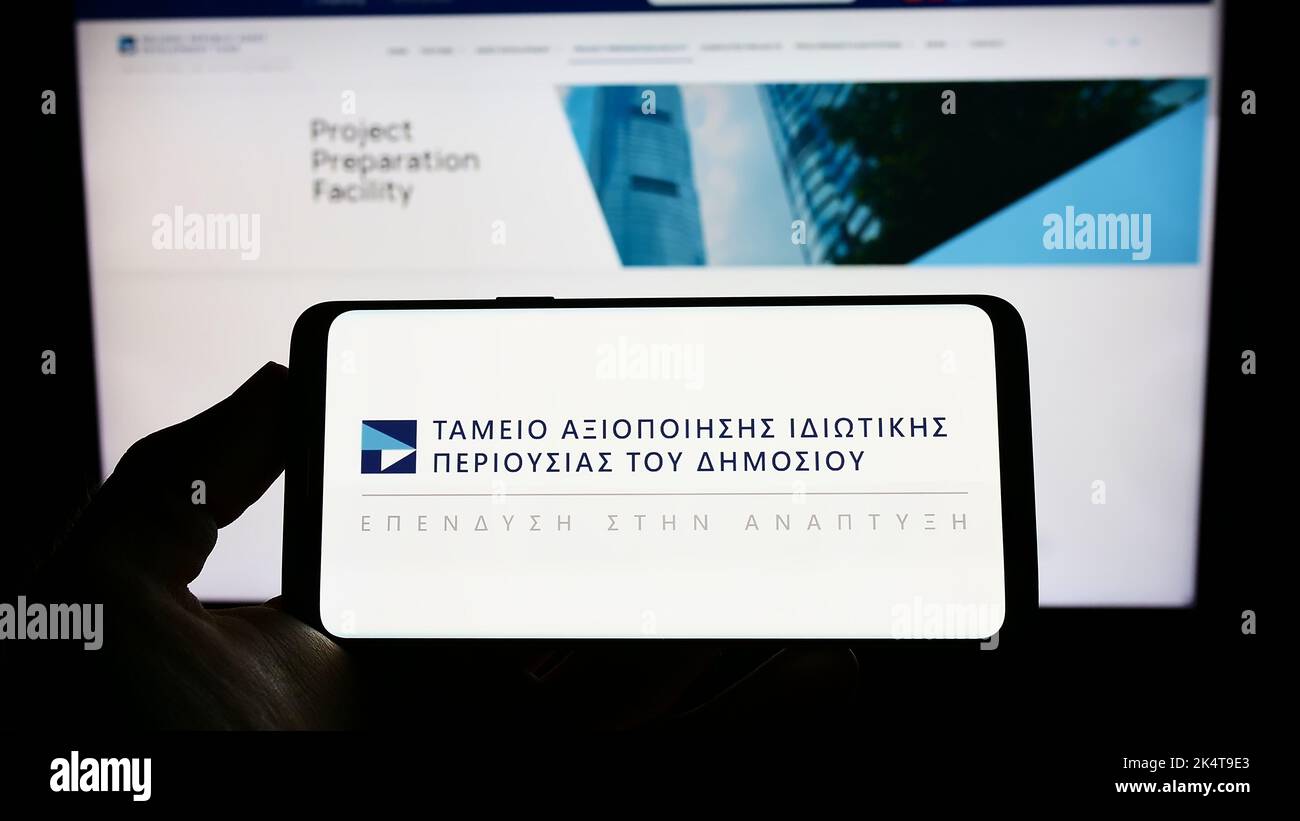 Persona che detiene un telefono cellulare con il logo del Fondo ellenico per lo sviluppo delle risorse (HRADF) sullo schermo davanti alla pagina web. Messa a fuoco sul display del telefono. Foto Stock