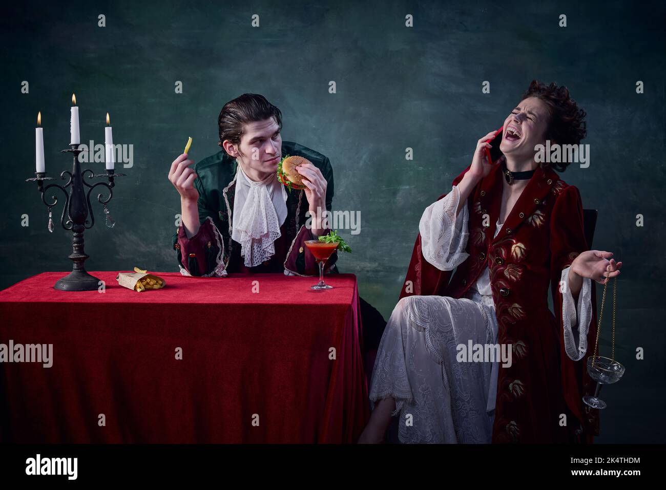 Ritratto di uomo e donna a immagine di vampiri medievali su sfondo verde scuro. Uomo che mangia hamburger, donna che parla al telefono Foto Stock