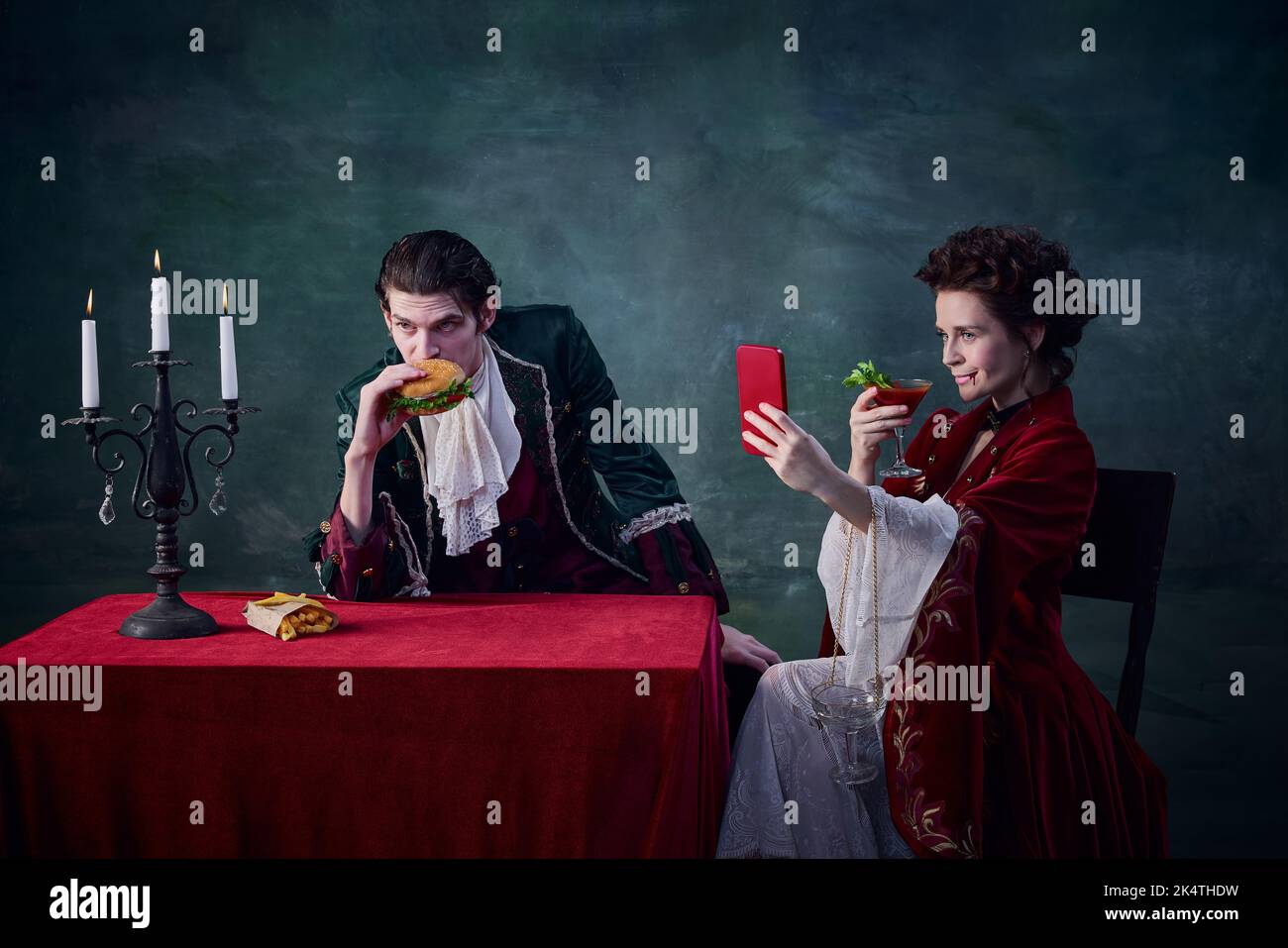 Ritratto di uomo e donna a immagine di vampiri medievali su sfondo verde scuro. Uomo che mangia hamburger, donna che beve e che prende selfie Foto Stock