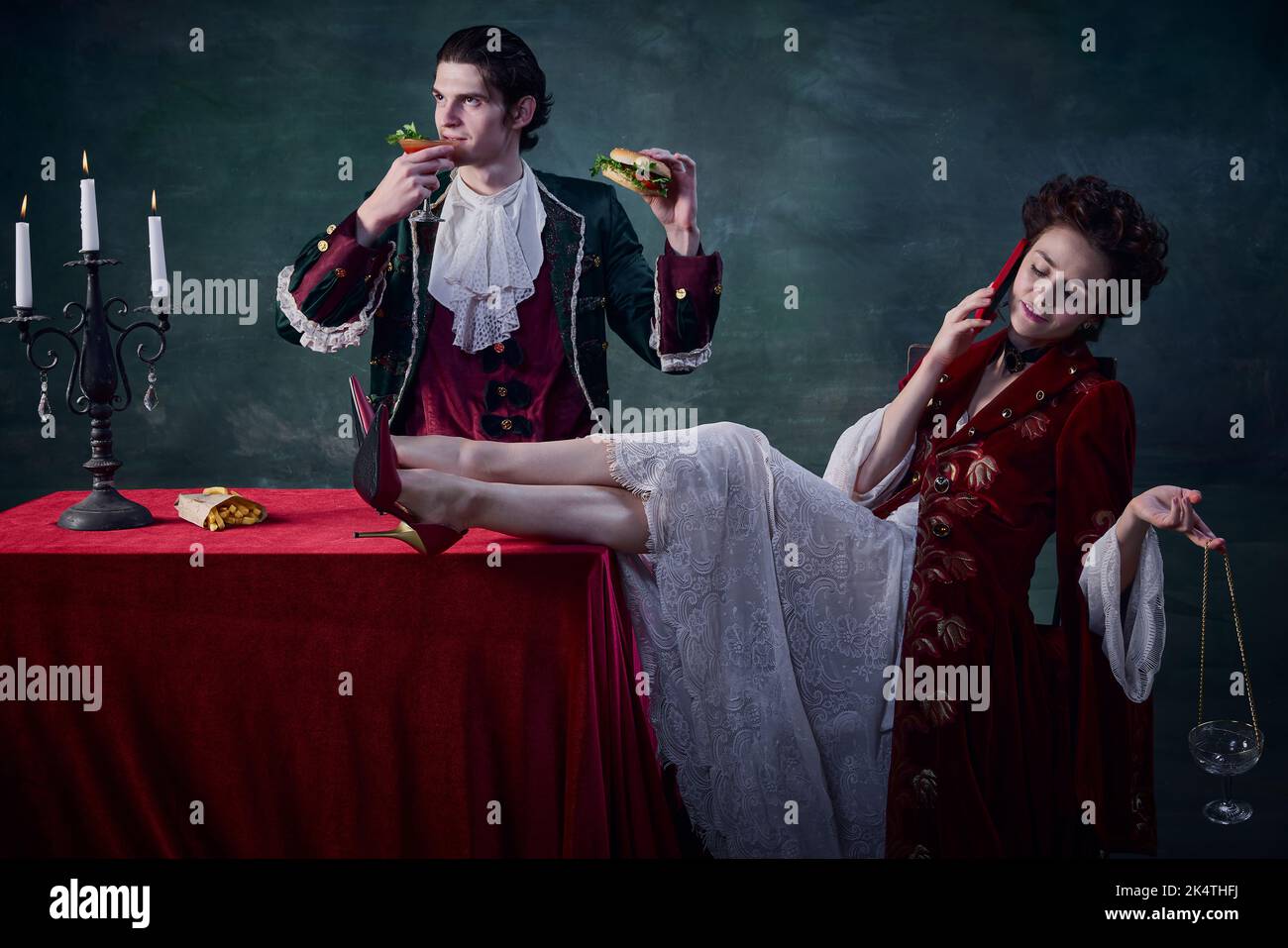 Ritratto di uomo e donna a immagine di vampiri medievali su sfondo verde scuro. Uomo che mangia hamburger e beve sanguinosa maria, donna che parla al telefono Foto Stock