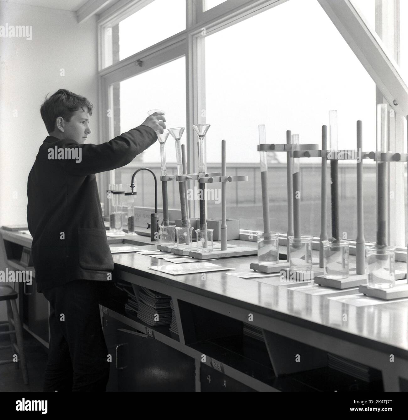 1964, storico, su una panchina vicino a una grande finestra, un allievo di scuola maschile in un laboratorio scientifico di una scuola secondaria che utilizza provette e contenitori di vetro per sperimentare e analizzare diversi liquidi, Inghilterra, Regno Unito. Foto Stock