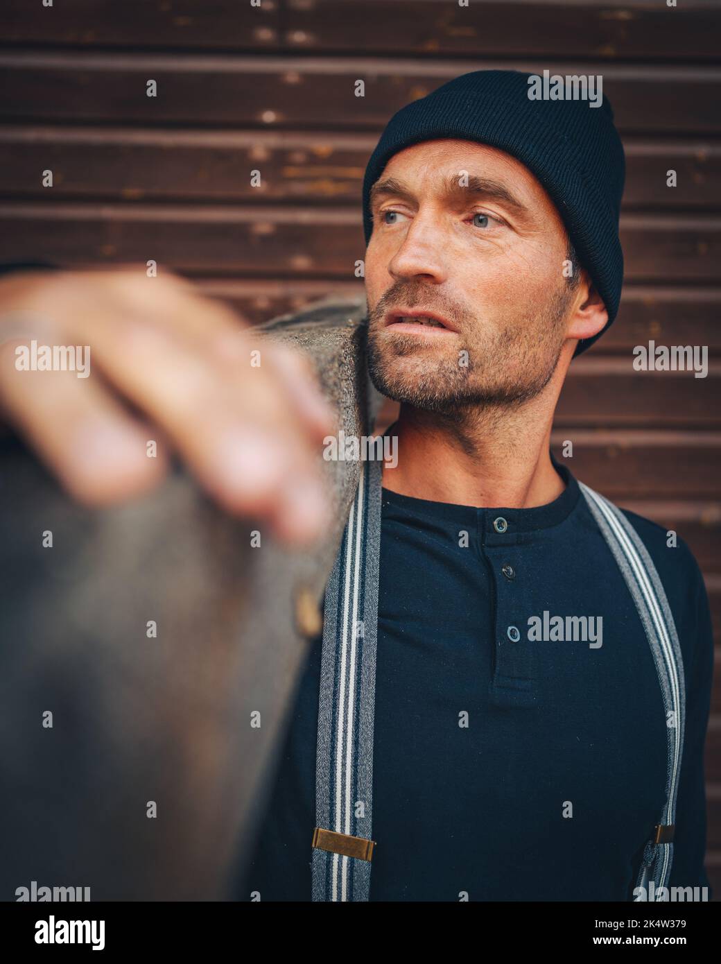 Buon uomo di lavoro osservante con il cappuccio e i sospensori sta trasportando una tavola di legno Foto Stock