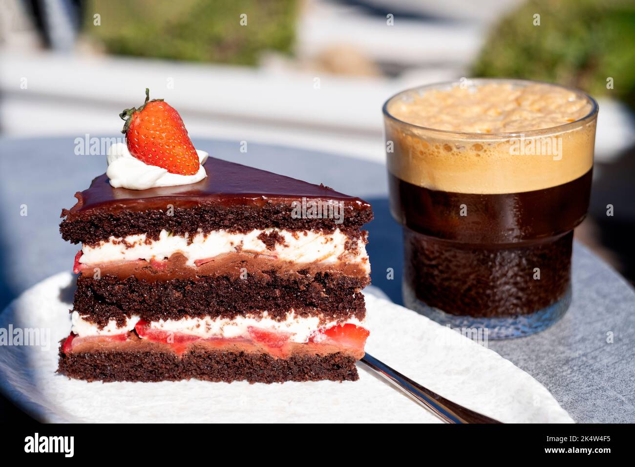 Una fetta di torta al cioccolato alla fragola di diavoli freschi servita su un tavolo da caffè all'aperto. la torta di panna fresca viene servita con un bicchiere di caffè ghiacciato Foto Stock