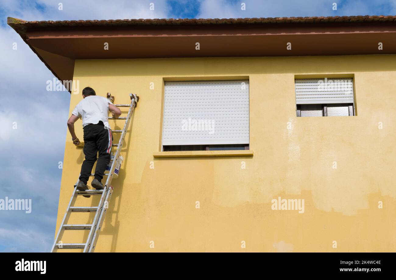 Vista posteriore di un uomo su una scala dipingendo una casa gialla, Spagna Foto Stock