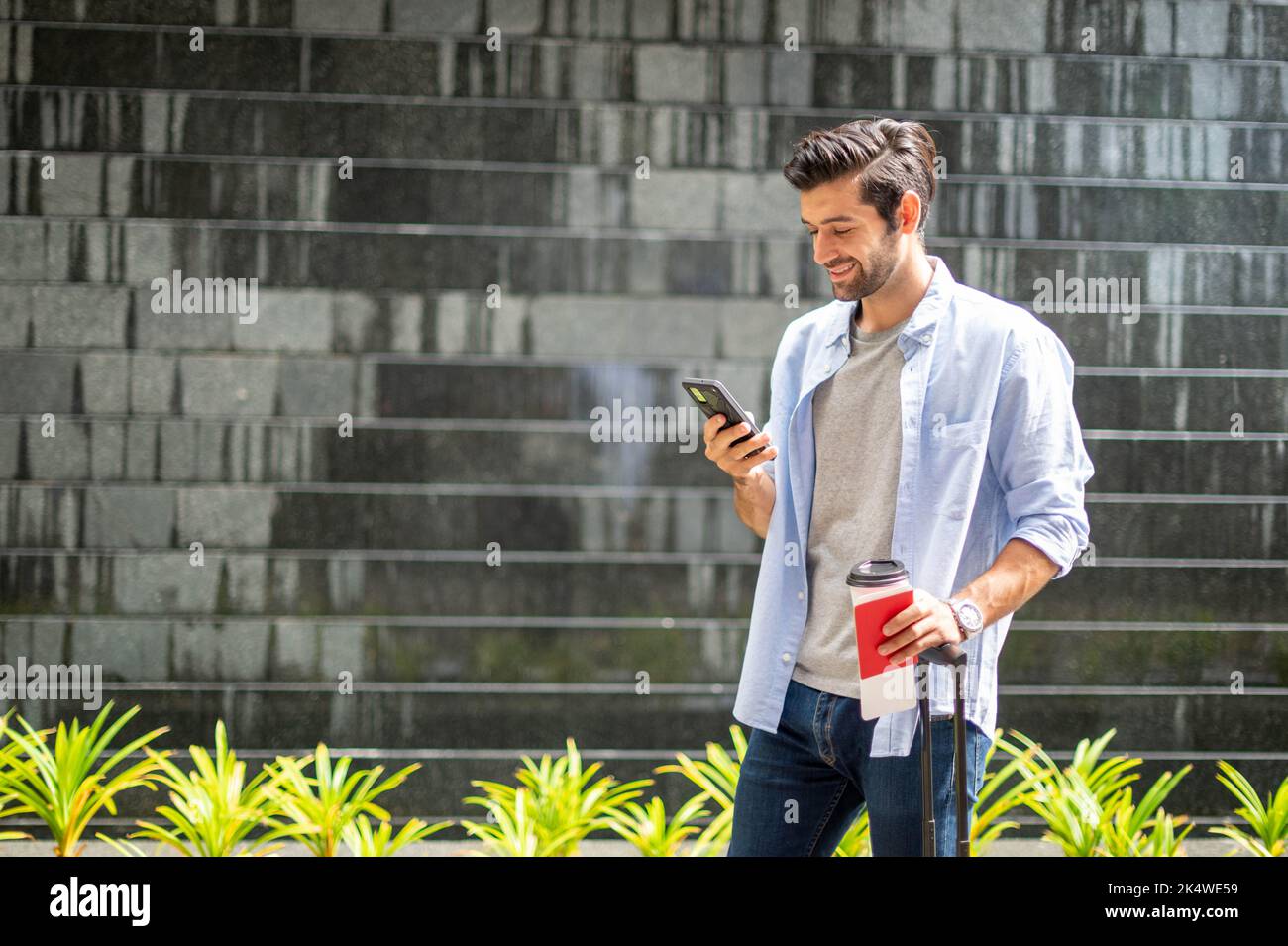 Giovane uomo caucasico con uno smartphone che tiene una tazza di caffè e tira la valigia in attesa che il suo amico si sposti insieme. Foto Stock