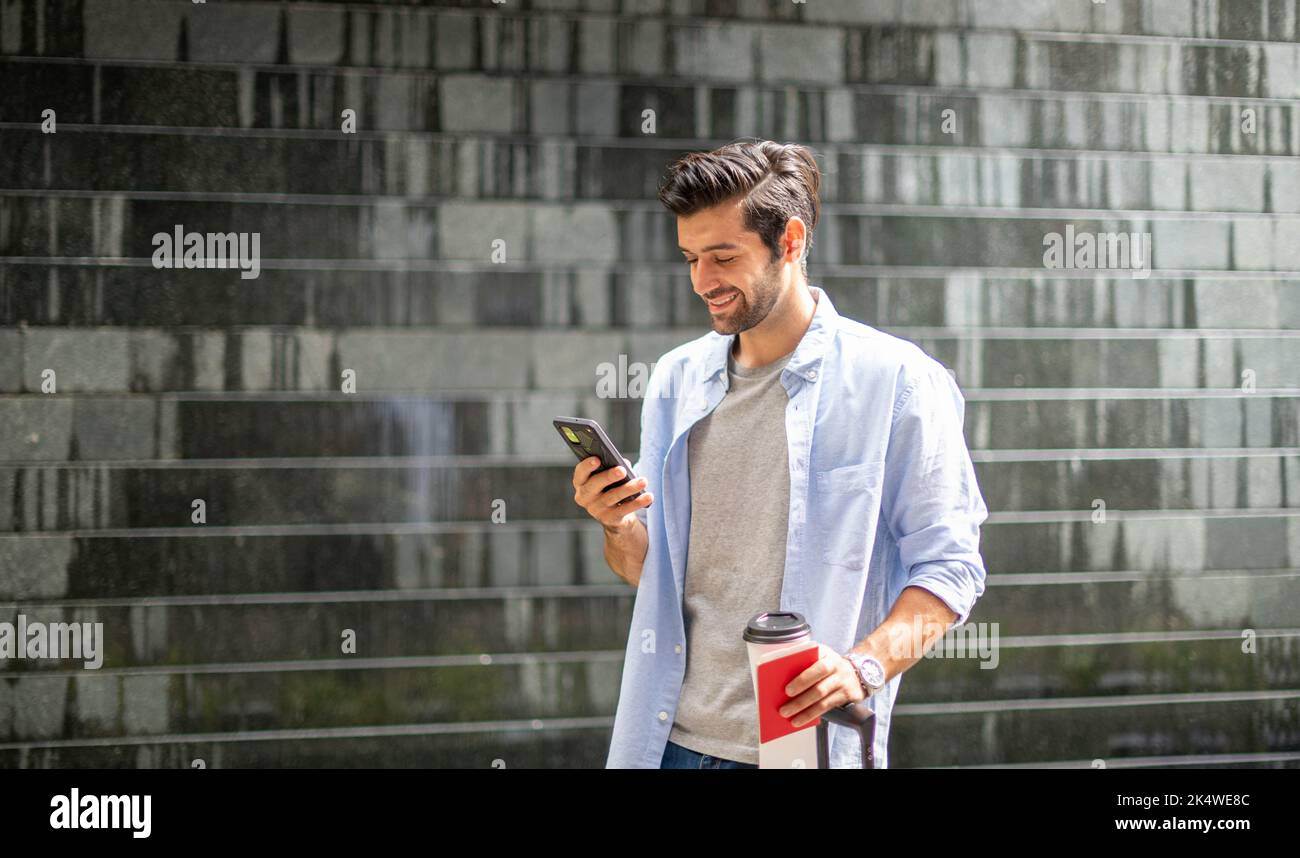 Giovane uomo caucasico con uno smartphone che tiene una tazza di caffè e tira la valigia in attesa che il suo amico si sposti insieme. Foto Stock