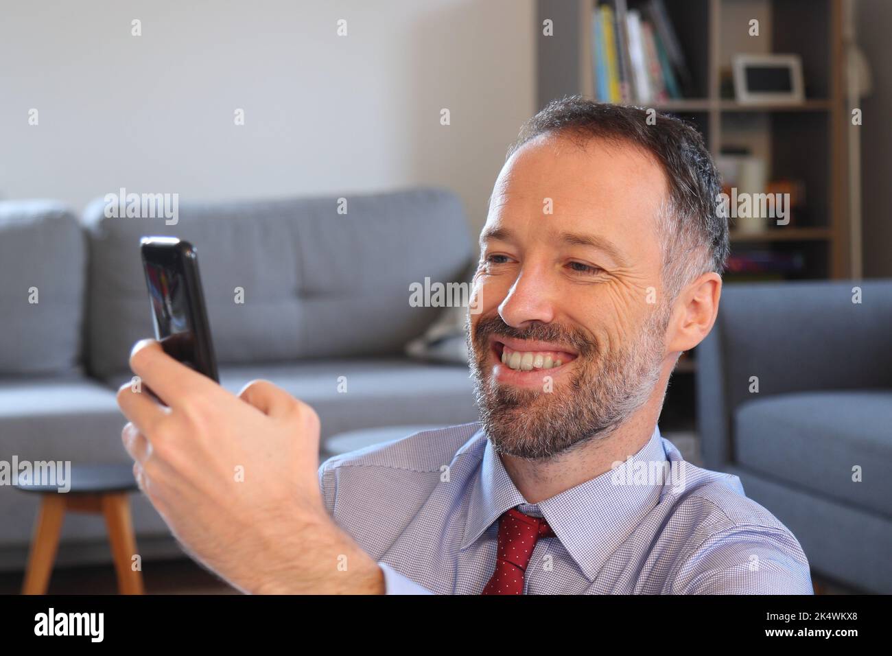 Uomo felice che usa lo smartphone a casa. Uomo portatore che indossa una maglietta da lavoro. Uomo caucasico adulto di 30s o 40s anni. Foto Stock