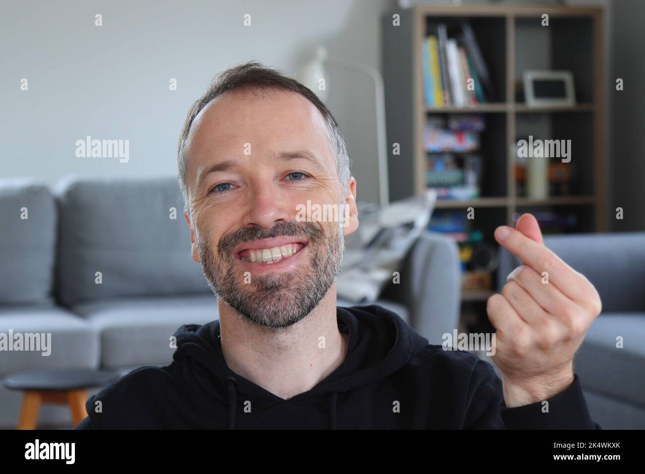 Uomo sorridente e che mostra un gesto cardiaco con le dita in stile asiatico o coreano. Uomo bearded con cappuccio nero casual. Uomo caucasico adulto in 30s o 40s sorridente Foto Stock