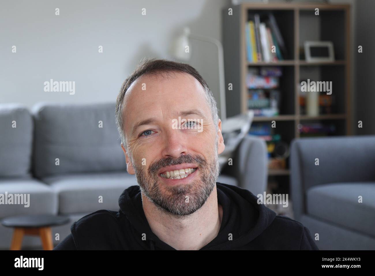Uomo sorridente a casa. Uomo bearded con cappuccio nero casual. Uomo caucasico adulto in 30s o 40s sorridente verso la macchina fotografica. Foto Stock