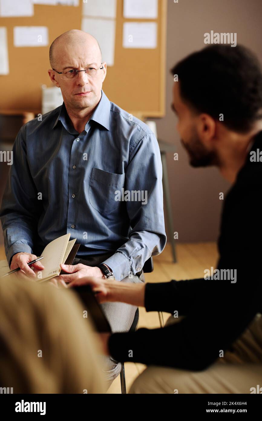 Consulente maturo fiducioso che guarda uno dei pazienti con disturbo post traumatico mentre lo ascolta durante la sessione Foto Stock