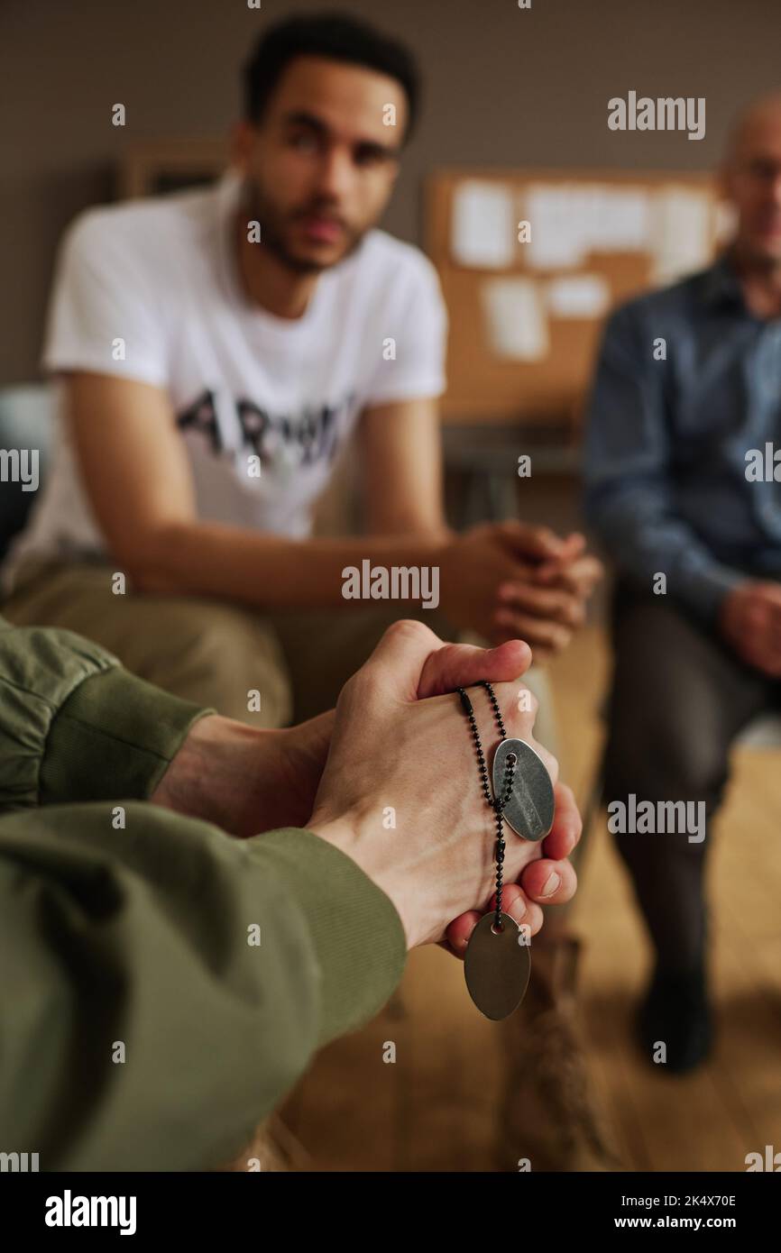 Mani di un giovane uomo con sindrome post traumatica catena di mantenimento con due medaglioni metallici mentre si siede tra gli altri assistenti Foto Stock