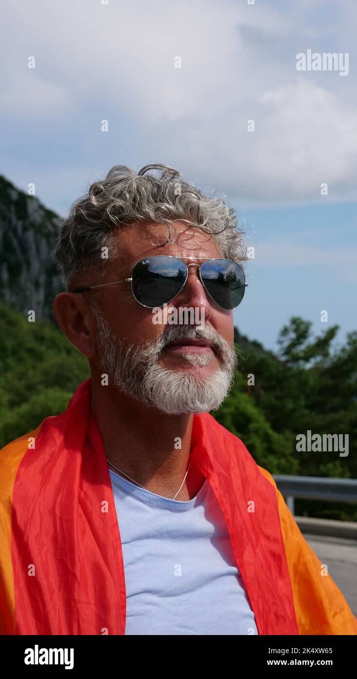 Ritratto di una bisessualità dell'uomo caucasico anziano dai capelli grigi con barba e occhiali da sole con bandiera arcobaleno LGBT in montagna. Celebra il mese dell'orgoglio, il giorno della bandiera dell'arcobaleno Foto Stock