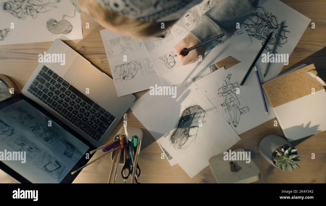 Vista dall'alto di una giovane donna che disegna i disegni a matita. Lavorare su uno storyboard di un fumetto in uno studio di design. Tavolo da lavoro di design con un computer portatile e barattolo fisso. Foto Stock