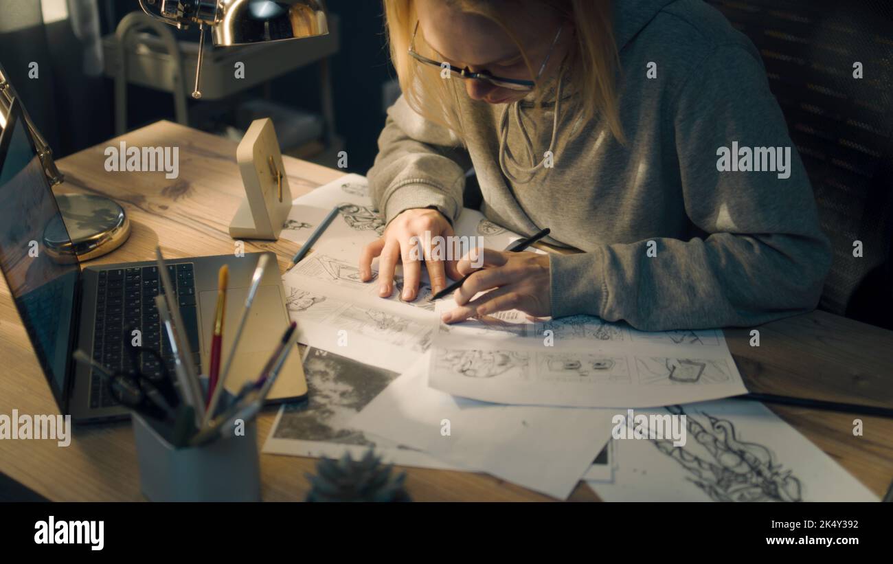 Una giovane donna lavora a uno storyboard in uno studio di design a casa. Un computer portatile e un vasetto fisso sul tavolo. La donna disegna gli schizzi come roadmap per il video. Pre-produzione. Foto Stock