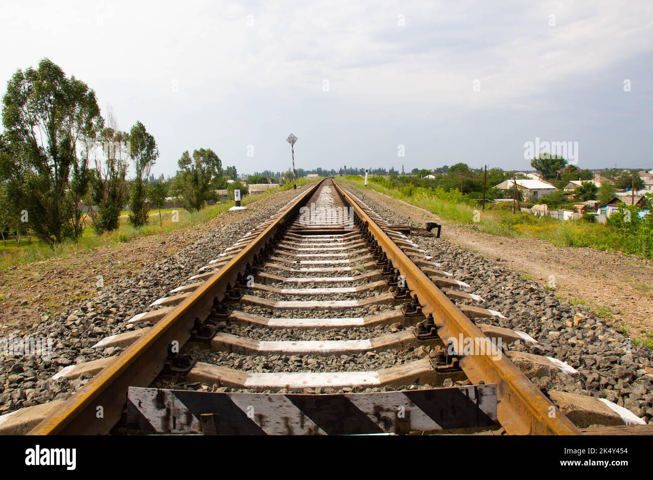 Ferrovia sopra l'orizzonte. Rotaie ferroviarie e traversine in cemento sullo sfondo di un'area rurale con case a un piano, alberi e un cielo blu Foto Stock