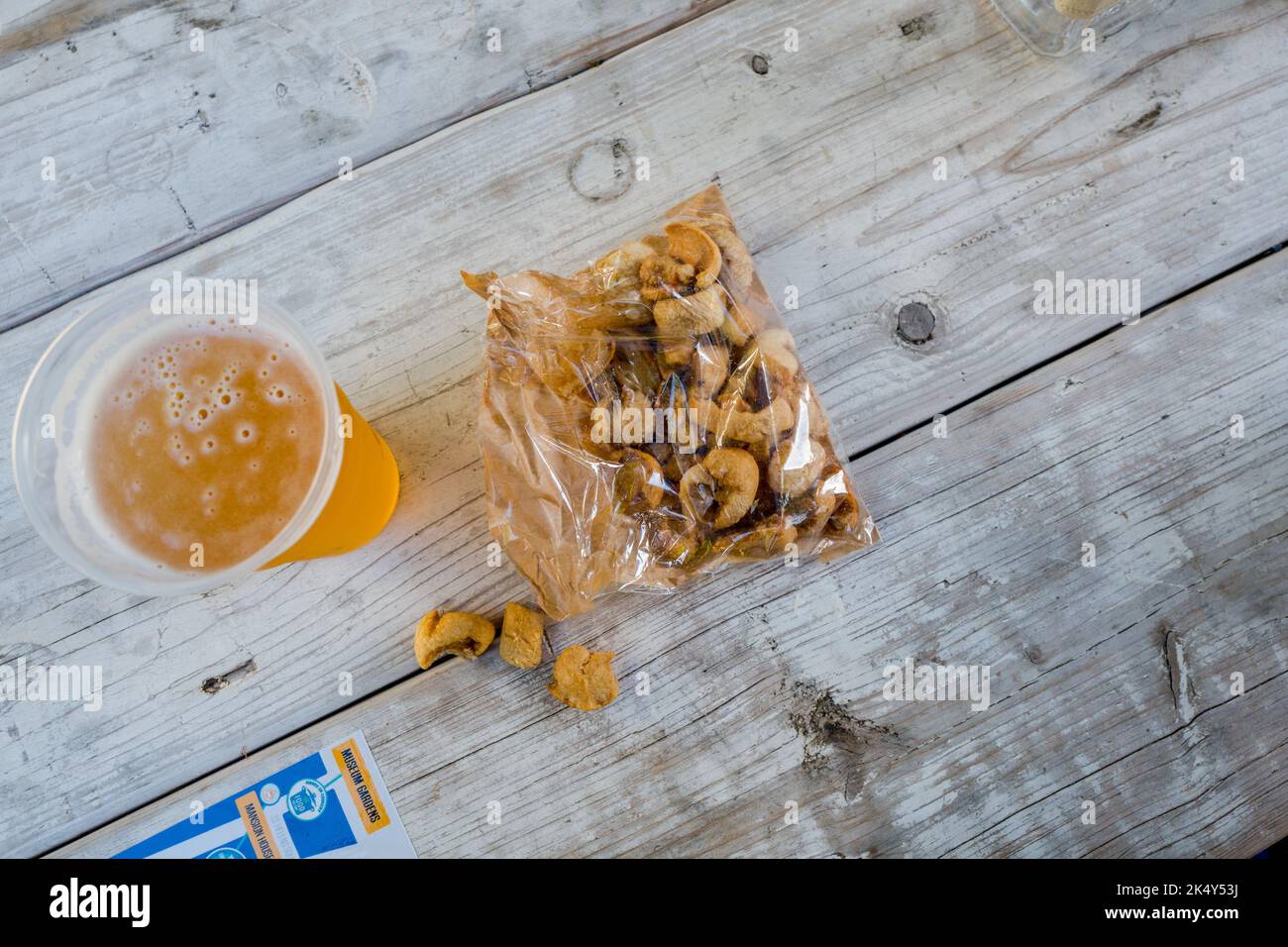 Una pinta di birra in un contenitore di plastica e un sacchetto aperto di cracklings o graffiature di maiale all'esterno su una panca di legno. Foto Stock